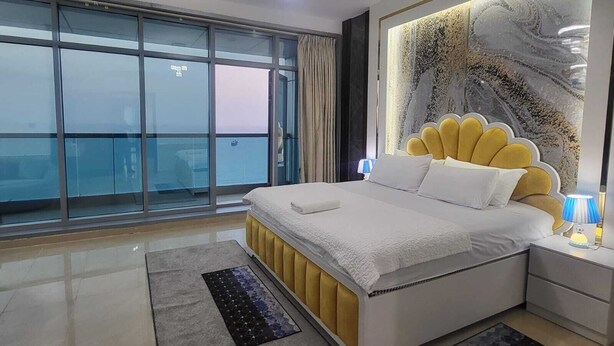 Une chambre moderne dotée d’un grand lit, offrant une vue sur la mer grâce à de grandes fenêtres du sol au plafond, et agrémentée d’un panneau décoratif mural.