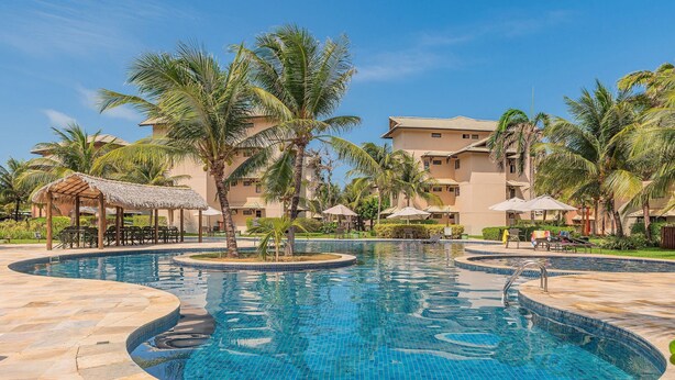 A swimming pool with a curved design, surrounded by palm trees and a building with multiple balconies. - Beach Place #202 - Seafront by Carpediem (Aquiraz)