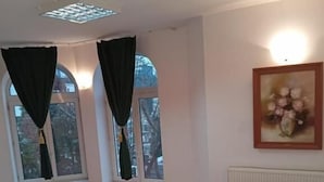 Quarto de hotel com uma cama, duas janelas com cortinas, um radiador e um quadro na parede.