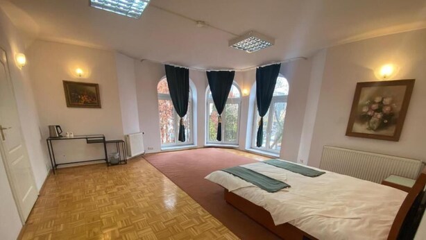 Quarto com piso deparquet de madeira, cama com manta verde, mesa de cabeceira e radiador. Possui duas janelas com cortinas, um quadro na parede e uma pequena mesa com um chaleira.