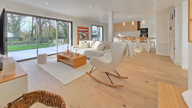 A modern living room with a large sliding glass door, a white sofa, a rocking chair, and a dining area with wooden chairs and a table. - New Home in Gulf of Morbihan & Vannes, Mahelia (Le Hézo)