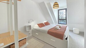 A modern bedroom with a sloped ceiling, a bed with a white and brown bedspread, a hanging light fixture, and a window with a view of trees. - New Home in Gulf of Morbihan & Vannes, Mahelia (Le Hézo)