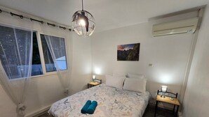 Ett sovrum med en säng, två sängbord, en hängande lampa, ett fönster med tunna gardiner och en väggmonterad luftkonditioneringsanläggning.