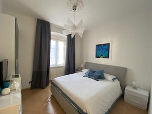 A bedroom with a large bed, a nightstand, a ceiling fan, a window with curtains, and a painting on the wall. - Câ Giâna apartment 5 minutes from the beaches (Lavagna)