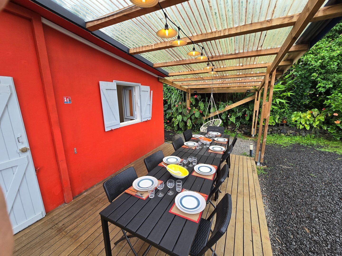 Un área de comedor al aire libre con una pérgola de madera, una pared roja y una mesa dispuesta para cuatro personas.