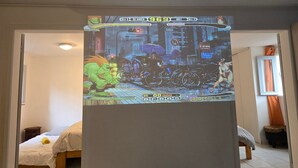 Ein Zimmer mit einem Bett, einem kleinen Tisch und einem an der Wand befestigten Bildschirm, der ein Videospiel zeigt.