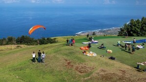 Ein grasbewachsener Hügel mit Menschen, einem Gleitschirmflieger und Blick auf das Meer.
