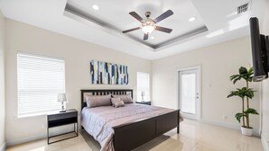 Une chambre à coucher comprenant un lit, des tables de chevet, un ventilateur de plafond, une grande fenêtre et une plante en pot.