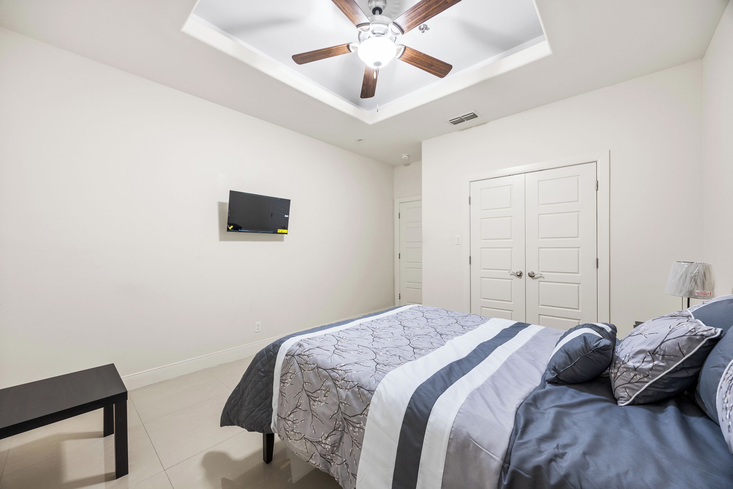 A bedroom with a bed, a ceiling fan, a TV mounted on the wall, and a door.