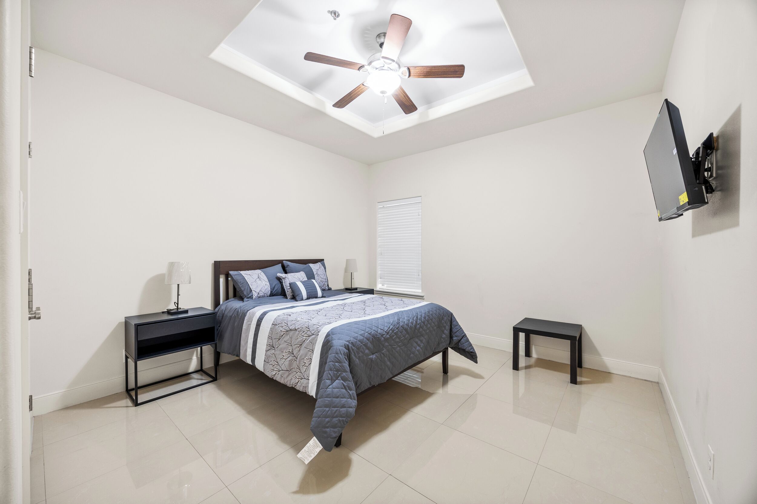 A bedroom with a bed, nightstands, a ceiling fan, a TV, and a window with blinds.