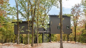 A modern treehouse with a staircase leading up to a platform, surrounded by trees and a gravel ground. - The Sun at In the Trees Luxury Resort🌳, Mountaintop, Hot Tub, Fireplace, Full Kitchen (Hot Springs National Park)