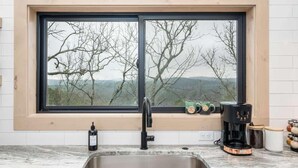 A kitchen with a large window, a stainless steel sink, a black faucet, a coffee maker, and a view of trees and hills. - The Sun at In the Trees Luxury Resort🌳, Mountaintop, Hot Tub, Fireplace, Full Kitchen (Hot Springs National Park)