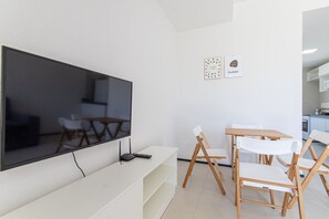 A modern living room with a flat-screen TV, wooden chairs, a white table, and a kitchen area in the background. - Exclusive Duplex at Beach Townhouse IV by Carpediem (Aquiraz)