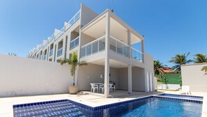 A modern building with a swimming pool, outdoor seating, and a clear blue sky. - Exclusive Duplex at Beach Townhouse IV by Carpediem (Aquiraz)