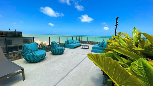 A rooftop terrace with blue seating, a view of the ocean, and tropical plants. - Garden Haus Flats - by Living (João Pessoa)