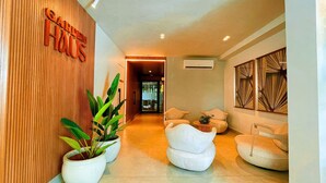 A modern hotel lobby with a wooden reception desk, potted plants, and a seating area with white armchairs and a small table. - Garden Haus Flats - by Living (João Pessoa)