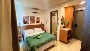 A hotel room with a bed, a wooden wardrobe, a bathroom with a sink and mirror, and a shower. - Garden Haus Flats - by Living (João Pessoa)