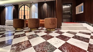 Um hall com piso de mármore quadriculado, área de estar com poltronas acolchoadas e uma grande janela com cortinas.