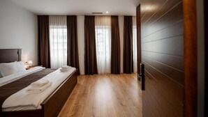 Quarto de hotel com piso de madeira, cama com lençóis brancos e janela com cortinas.