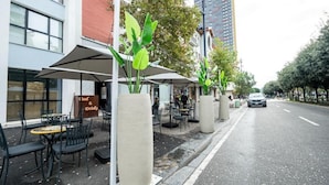Um café na rua com mesas ao ar livre, um letreiro anunciando comida e bebidas, e um edifício alto ao fundo.