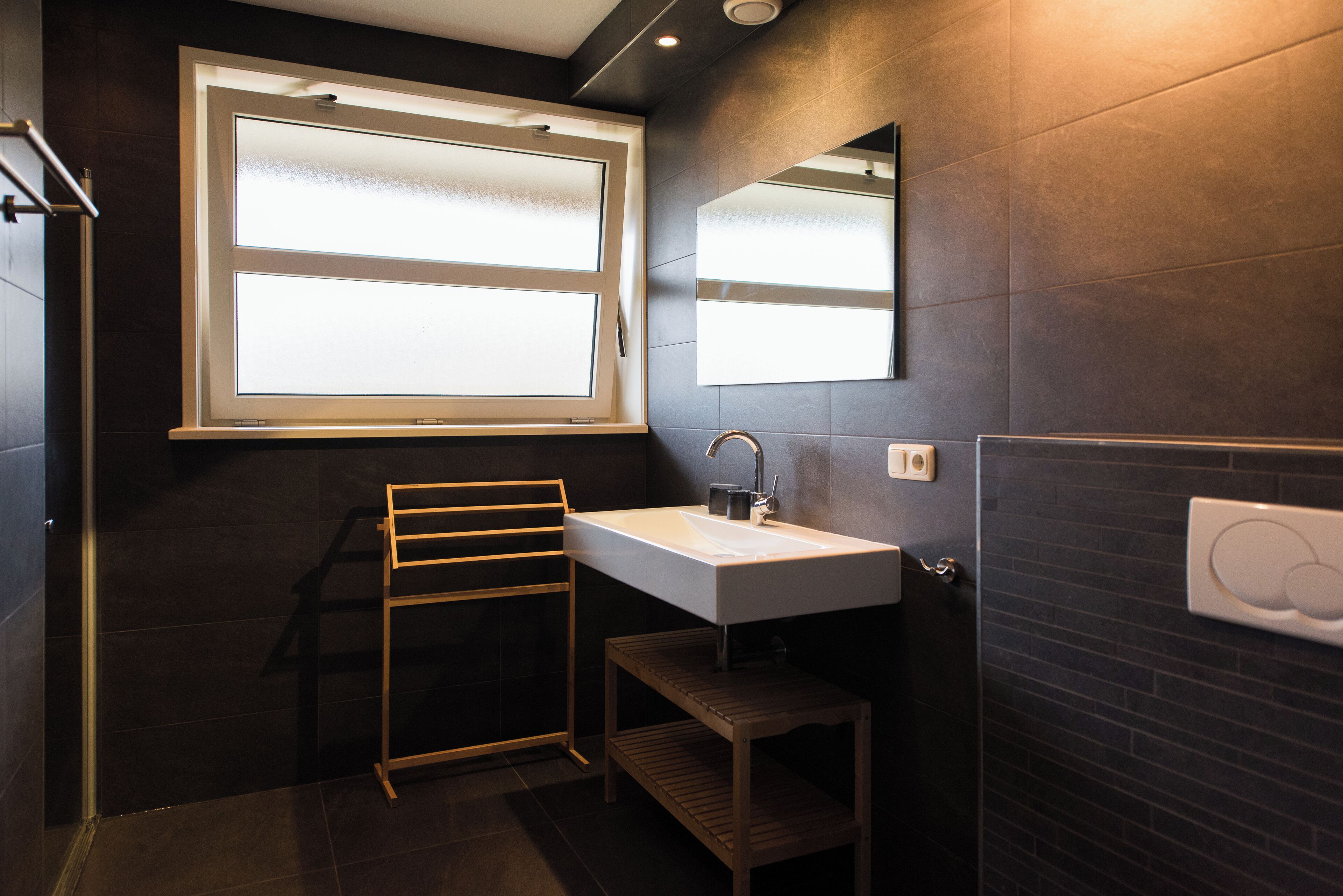 Un bagno moderno con una parete rivestita di piastrelle scure, un lavandino bianco, un portasciuga e una finestra con vetro smerigliato.