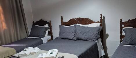 A hotel room with three beds, each with a wooden headboard, grey bedding, and a remote control on the bed in the center.
