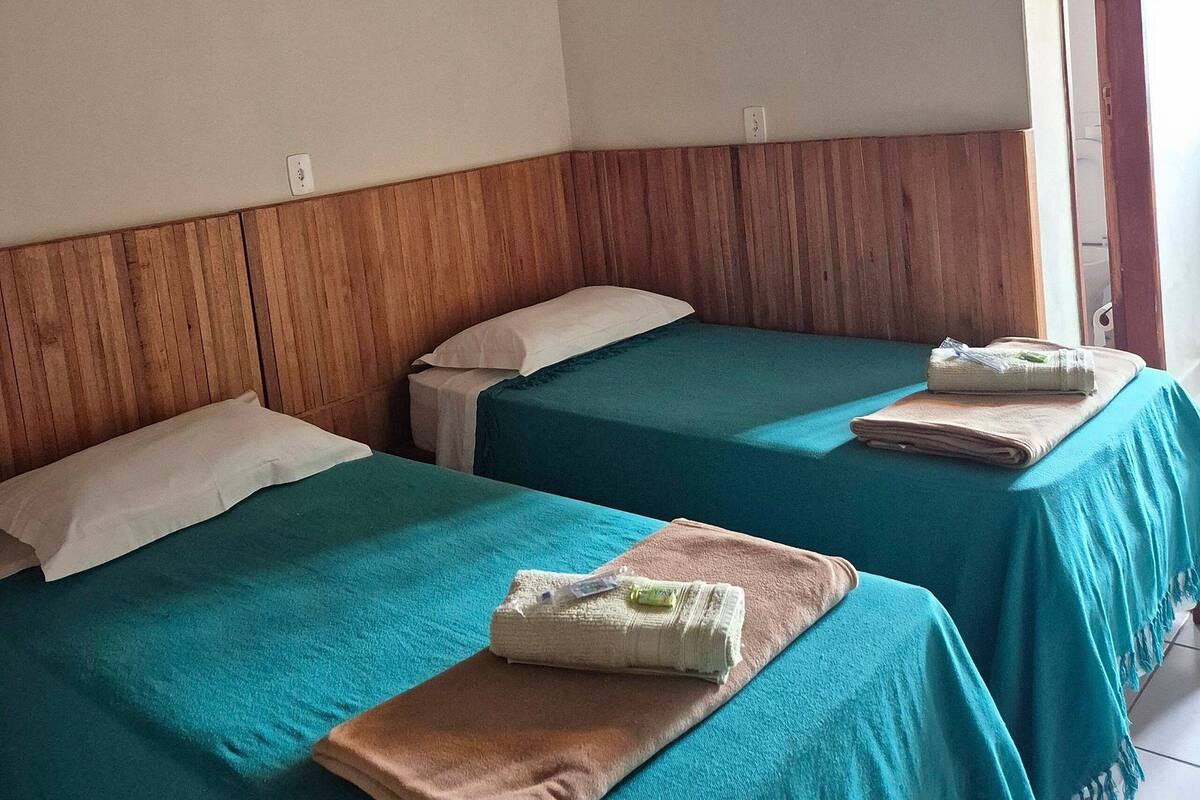 A room with two beds, each with a teal bedspread and a folded towel. The room has wooden paneling on the walls and a wooden ceiling.