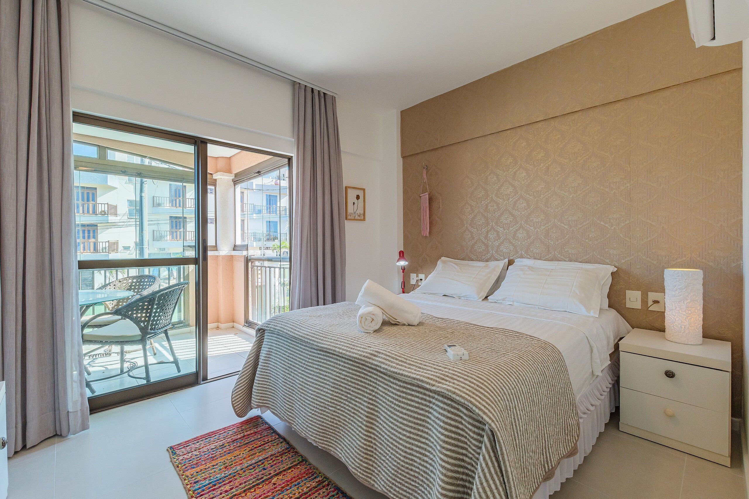 Un dormitorio con cama, mesita de noche, lámpara, ventana con cortinas y balcón con sillas.