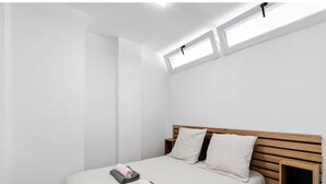 Quarto minimalista com uma cama, duas almofadas, uma mesinha de cabeceira e um claraboia.
