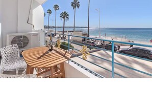 Uma varanda com mesa e cadeiras de madeira, vista para uma praia com palmeiras e céu claro.