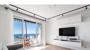 Uma sala de estar moderna com um sofá cinzento, uma TV de tela plana fixada numa parede branca e uma porta de vidro deslizante que dá para uma varanda com vista para o mar.
