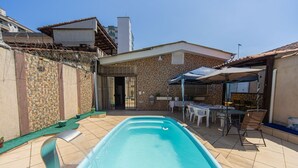 Une piscine avec une terrasse carrelée, un patio couvert avec des tables et des chaises, et un mur en pierre.