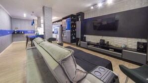 Un salon moderne avec un canapé, une table basse et un téléviseur fixé au mur. On aperçoit à l’arrière-plan un coin cuisine avec des placards et un four à micro-ondes.