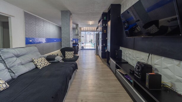 Un salon moderne avec un canapé, un téléviseur fixé au mur et un coin cuisine à l’arrière-plan.