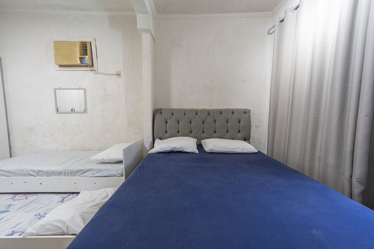 Quarto com duas camas de solteiro, uma com colcha azul e outra com padrão. Há um ar-condicionado fixado na parede e uma pequena janela com moldura de metal.