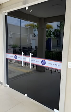A glass door entrance with a security barrier, a clear view of the outdoor area, and a building with a modern design. - 1403 Apto 2 Qtos Cabedelo João Pessoa (Cabedelo)