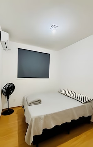 A small, single-bed room with a white bedspread, a black and white striped pillow, a standing fan, and a ceiling-mounted air conditioning unit. - 1403 Apto 2 Qtos Cabedelo João Pessoa (Cabedelo)