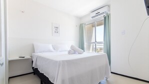Ein ordentlich bezogenes Bett mit weißen Leinen, ein Nachttisch, eine Wand-Klimaanlage und ein Fenster mit Vorhängen.