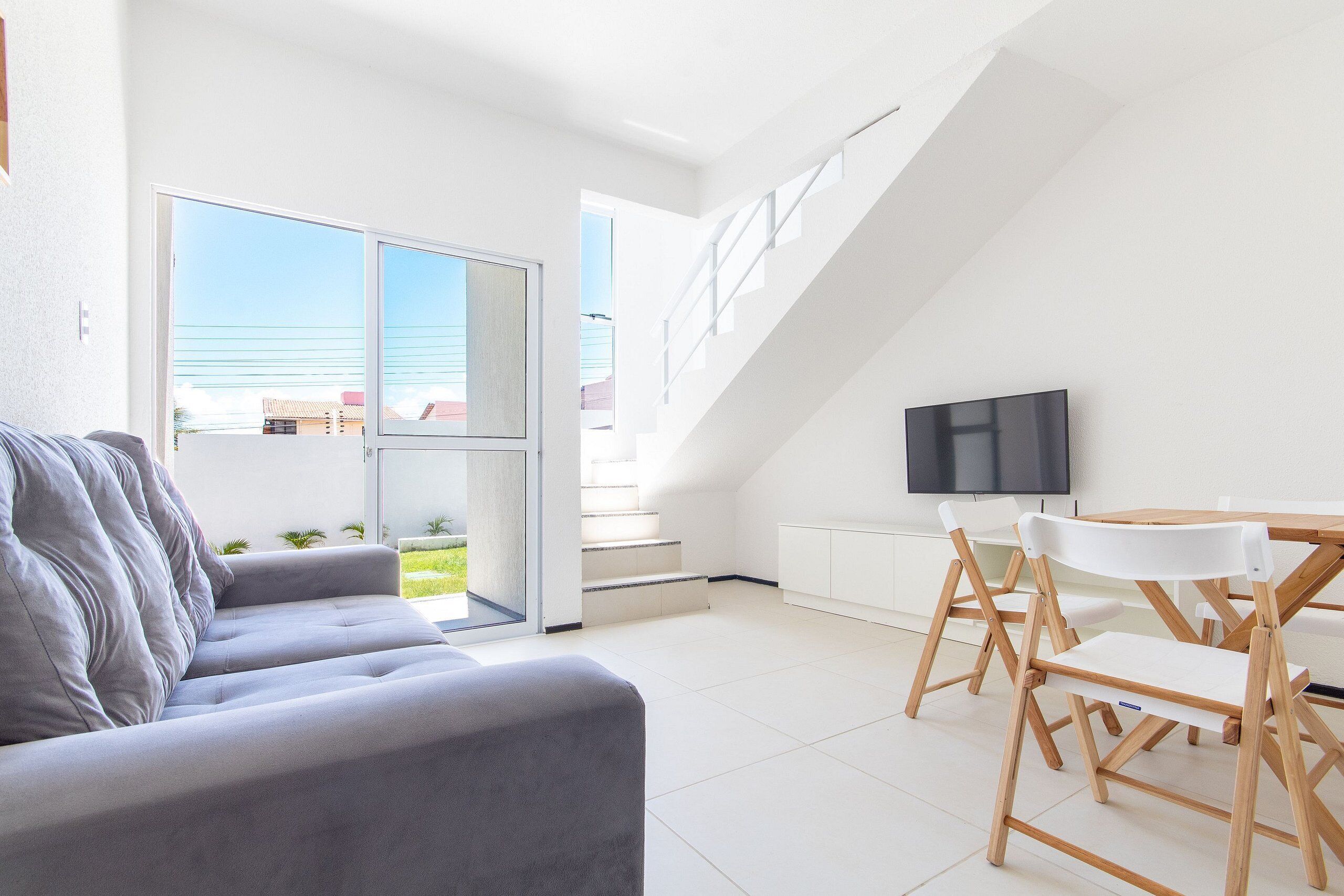 Ein modernes Wohnzimmer mit einem grauen Sofa, einem Esstisch mit Stühlen, einem Fernseher und einer Treppe, die zu einer oberen Etage führt.