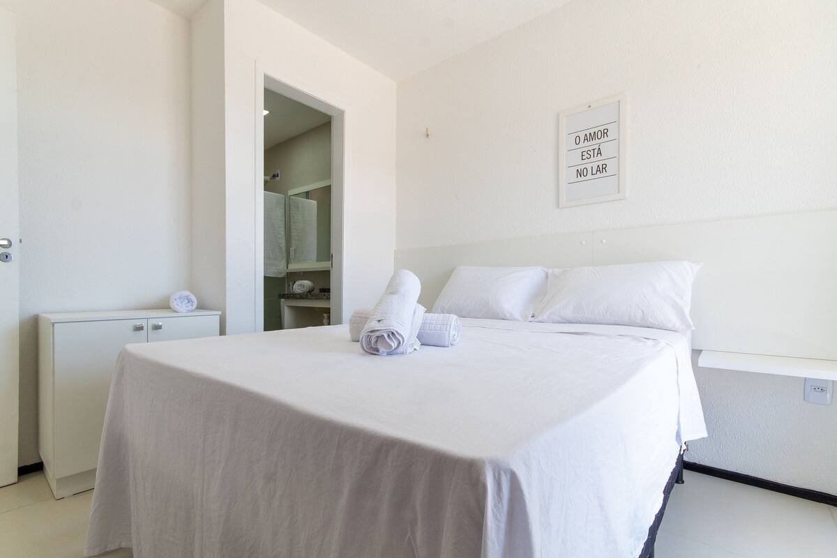 A neatly made bed with white linens and towels, a bedside table, a small cabinet, and a wall-mounted sign with text.
