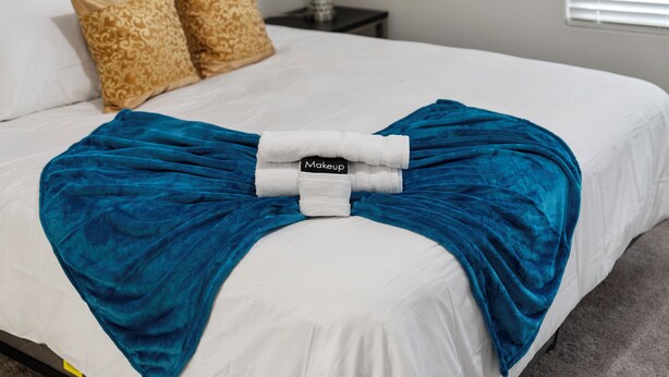 Een net opgemaakt bed met een witte deken, twee kussens met een patroon, een blauwe plaid en een witte handdoek met de tekst 'Makeup'.