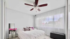 Une chambre à coucher comprenant un lit, un ventilateur de plafond, une fenêtre avec des rideaux légers, une table de chevet et un petit banc.