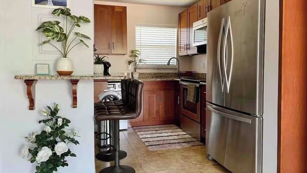Une cuisine avec des meubles de rangement en bois, un réfrigérateur en acier inoxydable, un four à micro-ondes et un évier. On y trouve deux plantes en pot et un tabouret de bar.