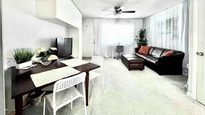 Un salon moderne comprenant un coin repas, un canapé en cuir noir, un ventilateur de plafond et une grande fenêtre avec des rideaux.
