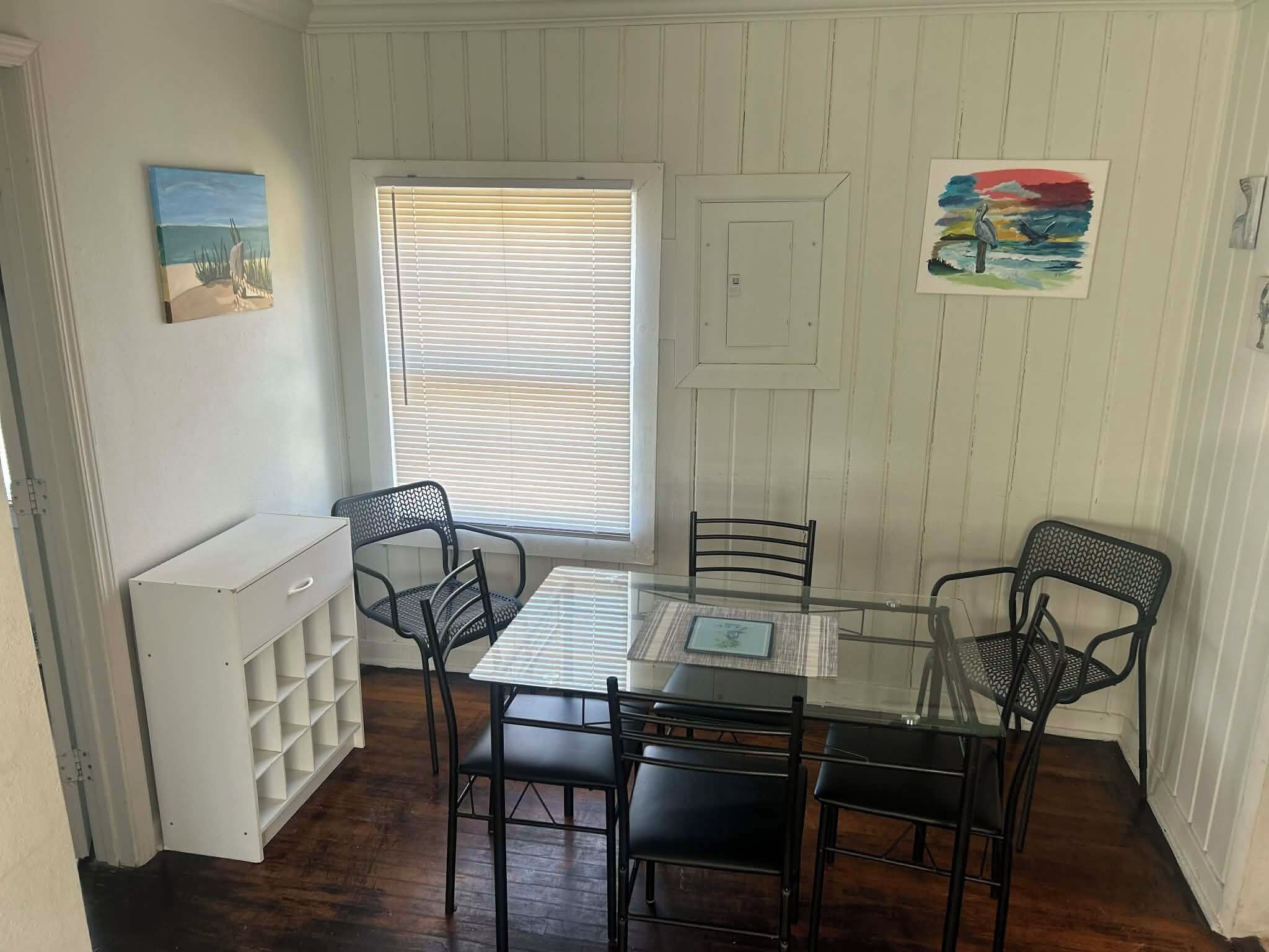 A dining area with a glass table and four chairs, a white radiator, and a window with blinds.