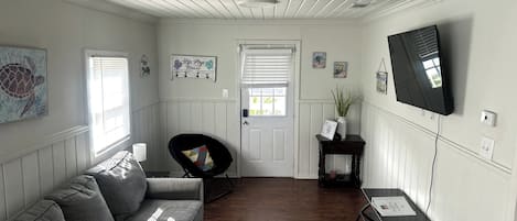 A living room with a grey sofa, a black chair, a small table with a vase, a mounted TV, and a door with a window.