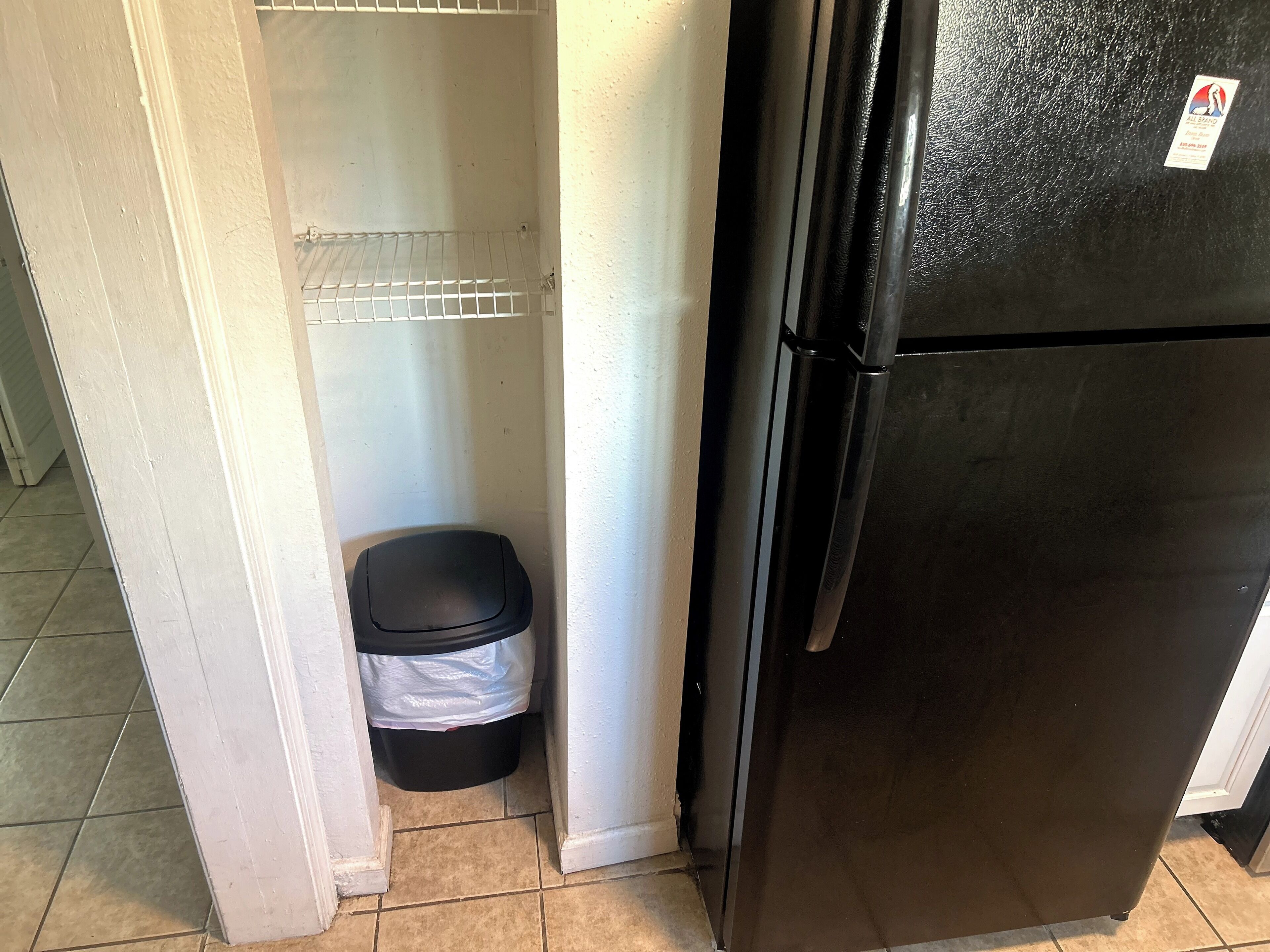 A small kitchen with a black refrigerator, a trash bin, and a white pantry.