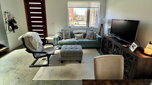 A living room with a sofa, ottoman, chair, TV, and a window with a view of buildings. - Home Base for World Cup (Kansas City)