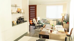 A modern living room with a sofa, coffee table, and dining area. There is a kitchen area with a coffee machine and shelves. A large window provides natural light. - Home Base for World Cup (Kansas City)
