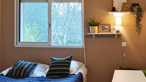 Ein Schlafzimmer mit Bett, Kissen, einem Fenster mit Jalousien, einem Regal mit einer Pflanze, einem gerahmten Bild und einer Nachttischlampe.